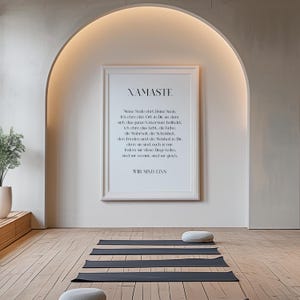 Puede incluir: Una impresi&oacute;n enmarcada con fondo blanco y texto negro que dice "NAMASTE" y una cita en alem&aacute;n. La impresi&oacute;n est&aacute; colgada en una pared blanca con una puerta arqueada encima. Hay cuatro esteras de yoga negras en el suelo de madera frente a la impresi&oacute;n.