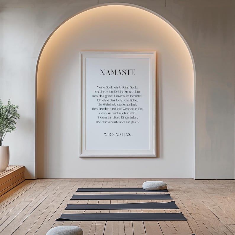 Puede incluir: Una impresi&oacute;n enmarcada con fondo blanco y texto negro que dice "NAMASTE" y una cita en alem&aacute;n. La impresi&oacute;n est&aacute; colgada en una pared blanca con una puerta arqueada encima. Hay cuatro esteras de yoga negras en el suelo de madera frente a la impresi&oacute;n.