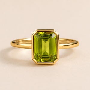 Puede incluir: Un anillo de oro con una gran piedra preciosa de color verde lima, de corte esmeralda rectangular, engastada en un bisel liso y pulido. La banda es fina, redondeada y está hecha de metal de oro amarillo pulido, con un diseño limpio.