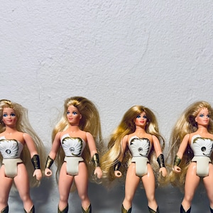 May include: Four vintage dolls with blonde hair and blue eyes. Each doll wears a white and gold outfit with a chest emblem, gold armbands, and boots. The dolls stand against a plain white background.