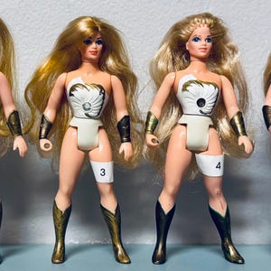 May include: Several vintage action figures of a female character with long blonde hair, wearing a white and gold outfit, and knee-high boots. The figures have a number on their leg.