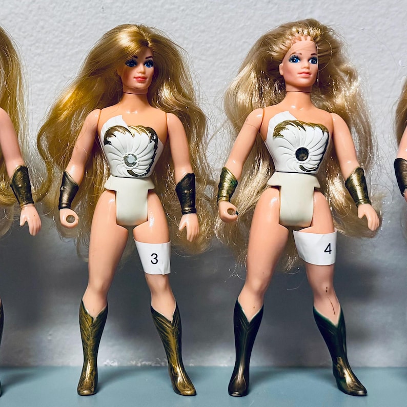 May include: Several vintage action figures of a female character with long blonde hair, wearing a white and gold outfit, and knee-high boots. The figures have a number on their leg.