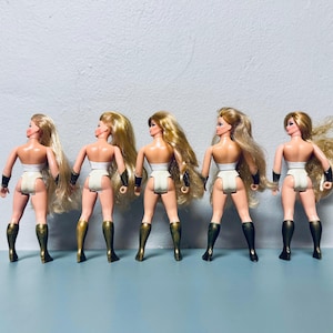 May include: Five vintage action figures of a female superhero, viewed from the back. Each doll has long blonde hair, a white bodysuit, gold and black armbands, and gold and black boots. The dolls are standing in a row.