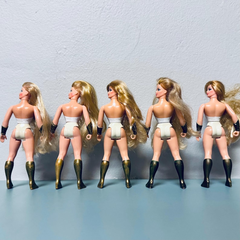 May include: Five vintage action figures of a female superhero, viewed from the back. Each doll has long blonde hair, a white bodysuit, gold and black armbands, and gold and black boots. The dolls are standing in a row.