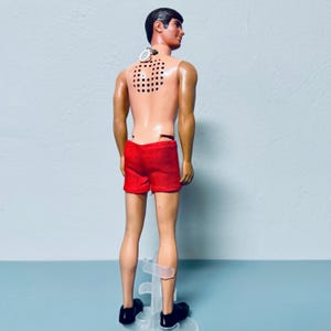 May include: A vintage doll with dark hair, wearing red shorts and black shoes. The doll has a grid pattern on its back and a small device attached to its neck. The doll is standing on a clear plastic stand.