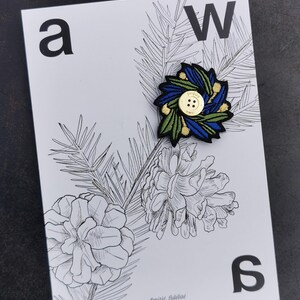 Pode incluir: Um patch floral bordado com detalhes azuis, verdes e dourados, centrado com um botão dourado. O patch está colocado num cartão branco com uma ilustração botânica a preto e branco e as letras "a", "W" e "s". O texto "Amitié, fidélité" também está presente.