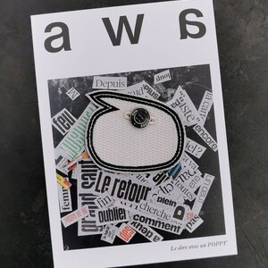Può includere: Una carta bianca con la scritta "aws" e una toppa a forma di fumetto in bianco e nero con un bottone. La toppa è su uno sfondo di ritagli di giornale. Il testo "Le dire avec un POPPY" è in basso.