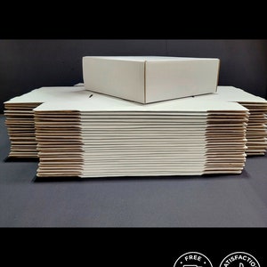 May include: A stack of flat, white cardboard shipping boxes with one assembled box resting on top. The boxes are constructed from plain, matte white paperboard with visible corrugated edges, designed for packaging and mailing various items.