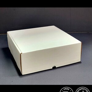 May include: A square white cardboard shipping box with a flat top and a front-facing tab closure. The box is constructed from plain white material and features clean, straight edges, presenting a simple and functional design for packaging items.