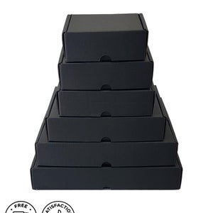 May include: A stack of six black cardboard boxes of varying sizes, arranged from largest at the bottom to smallest at the top. Each box has a lid with a small tab for opening. The boxes are on a white background. Two circular icons are in the lower left corner.
