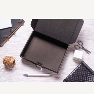 May include: An open black cardboard box with a textured interior sits on a light wood surface. A ball of twine, a white pen, scissors, a white cube, a black bubble mailer, and a notebook are arranged around the box, suggesting packaging supplies.