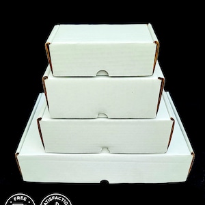 May include: A stack of four white rectangular cardboard shipping boxes arranged in a pyramid shape, with the smallest box on top and the largest on the bottom. Each box features a front-facing tab closure and a clean, matte white exterior finish.