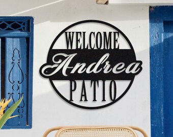 Könnte beinhalten: Ein rundes Schild in Schwarzweiß mit dem Text "Welcome Andrea Patio" hängt an einer weißen Wand. Ein Korbstuhl mit einem schwarz-weiß karierten Kissen steht vor einer blauen Tür.
