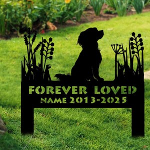 May include: Black metal memorial garden stake featuring a silhouette of a dog sitting amidst stylized plants. The text "FOREVER LOVED" and a name with dates are cut out of the metal. The stake is placed in a grassy area with colorful flowers in the background.