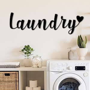Può includere: Una lavatrice bianca è posizionata accanto a un mobile contenitore bianco con cesti in vimini. La parola "Laundry" è visualizzata in corsivo nero sopra la macchina, con un simbolo a forma di cuore. Piante e oggetti decorativi completano la scena.