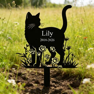 May include: Black metal cat memorial stake with the name "Lily" and dates 2010-2026. The cat silhouette is surrounded by a floral design with roses and grass. The stake is placed in a grassy field.