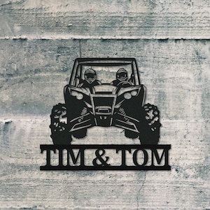 Könnte beinhalten: Schwarze Metallsilhouette eines Side-by-Side-Geländewagens mit zwei Figuren auf dem Fahrer- und Beifahrersitz. Das Fahrzeug hat eine schwarze Metallplakette darunter, auf der "TIM & TOM" steht.