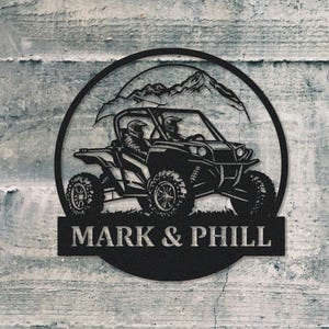 May include: Black metal silhouette of a two-seater off-road vehicle with a mountain backdrop. The vehicle is driving on a dirt road. The text "MARK & PHILL" is below the vehicle.