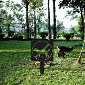 May include: Black metal sign with a white circle and a diagonal line through it. Inside the circle is a silhouette of a dog. The sign is on a black metal stake and is in a grassy area.