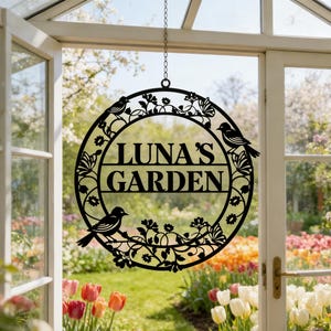 Puede incluir: Un letrero circular de metal negro para colgar con el texto LUNA'S GARDEN en el centro. El marco está decorado con intrincados recortes de pájaros, mariposas y enredaderas florales. El letrero está suspendido por una fina cadena.