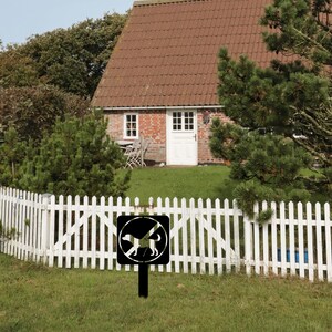 May include: A black metal sign with a white silhouette of a dog with a diagonal line through it. The sign is mounted on a black metal post and is in front of a white picket fence. The sign reads "No Dogs Allowed".