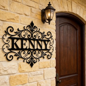 May include: Black metal sign with the name "KENNY" in bold letters, surrounded by ornate scrollwork. The sign is mounted on a light-colored stone wall, next to a dark wooden door and a vintage-style outdoor lamp.