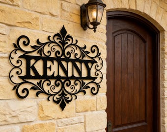 May include: Black metal sign with the name "KENNY" in bold letters, surrounded by ornate scrollwork. The sign is mounted on a light-colored stone wall, next to a dark wooden door and a vintage-style outdoor lamp.
