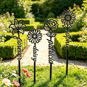 Könnte beinhalten: Vier schwarze Metall-Gartenstecker mit lasergeschnittenen Blumenmotiven an der Spitze. Jeder Stecker zeigt einen Namen in Schreibschrift: forever, daisy, patricia und sophie. Die Blumen umfassen eine Lilie, ein Gänseblümchen, eine Rose und eine Sonnenblume.