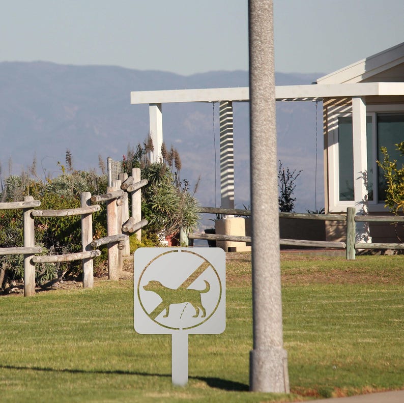 May include: A white metal sign with a black silhouette of a dog and a diagonal line through it. The sign reads "No Dogs" and is mounted on a white metal post. The sign is in front of a wooden fence and green grass.