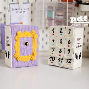 Może przedstawiać: Dwa pudełka na szydełku, jedno fioletowe z żółtą ramką, a drugie białe z czarnymi cyframi od 1 do 12. Oba pudełka mają napis "Oh My God" i odcisk dłoni.