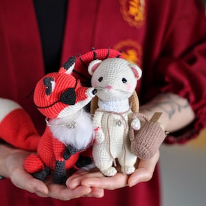 May include: A white crocheted mouse with a brown crocheted bag, and a red crocheted fox with a white and black tail, both wearing necklaces.