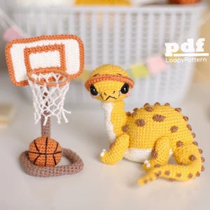 Puede incluir: Un gecko de crochet amarillo con manchas marrones jugando al baloncesto. El gecko lleva un gorro naranja de crochet. El aro de baloncesto es marrón con una red blanca y una pelota de baloncesto marrón.