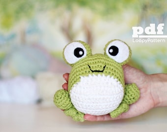 May include: Green crocheted frog amigurumi toy with white belly and large black eyes. The frog is being held in a hand.