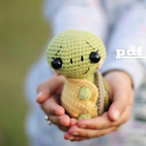Op de afbeelding: Een handgemaakte gehaakte schildpoppenpop in groen en geel. De schildpad heeft grote zwarte ogen, een lachende mond en een geel vest. De pop wordt in komvormige handen gehouden, met de tekst "pdf LoopyPattern" zichtbaar.