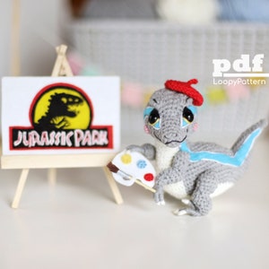 Könnte beinhalten: Ein grauer gehäkelter Dinosaurier mit einem roten Barett hält eine Farbpalette und einen Pinsel. Der Dinosaurier sitzt vor einem kleinen Staffelei mit einem Jurassic Park-Logo.
