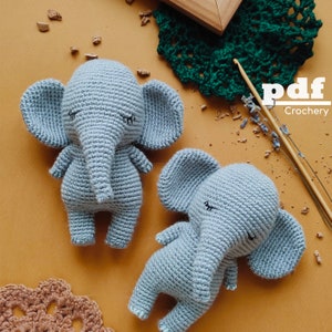 May include: Two crocheted gray elephant amigurumi toys. The larger elephant is standing upright, while the smaller elephant is lying down. Both elephants have closed eyes and a light gray color.