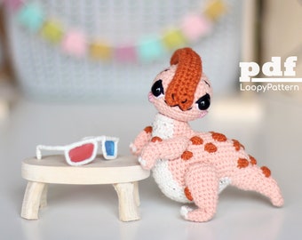 May include: A handmade crochet dinosaur in pink, white, and orange, with dark eyes and orange spots. It is next to a small wooden table with 3D glasses. The image includes the text "pdf" and "LoopyPattern".