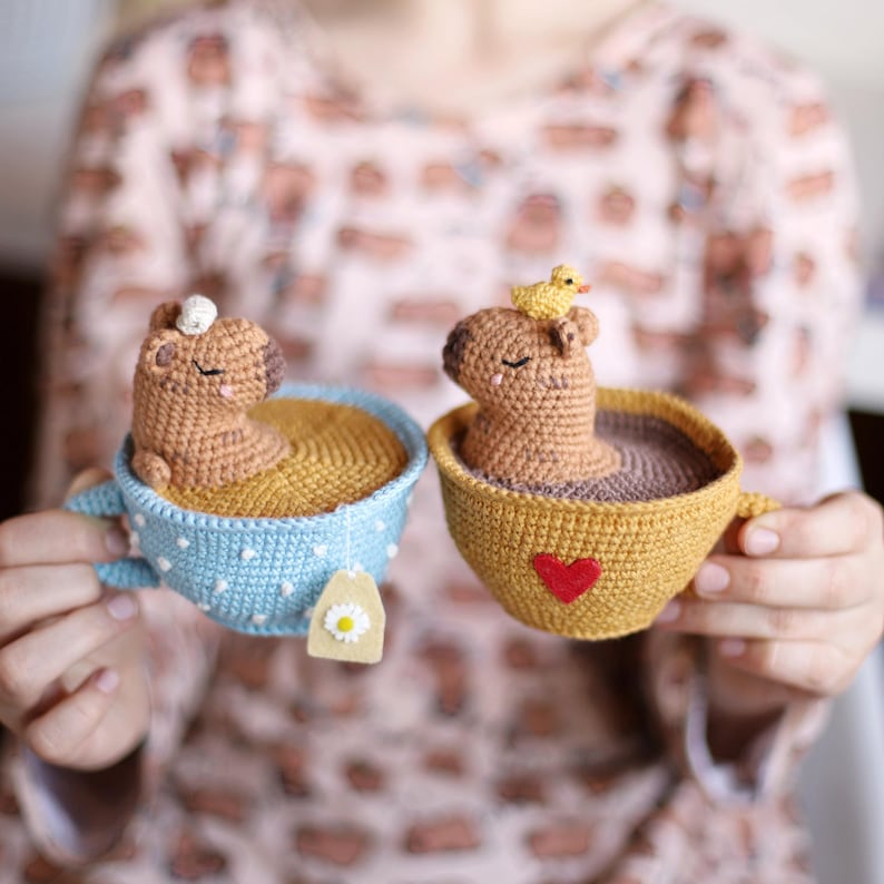 May include: Two crocheted stuffed animals, resembling capybaras, are sitting in teacups. One capybara is in a blue teacup with white polka dots and a tea bag tag. The other capybara is in a yellow teacup with a red heart and a small yellow crocheted duck on its head.