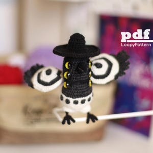 May include: A handmade black and white crochet bird with a black hat and multiple yellow eyes. The bird has white and black striped wings and is perched on a white stick. The image includes the text "pdf LoopyPattern".