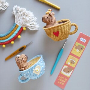 May include: Two crocheted capybara amigurumi toys sitting in teacups. One capybara is in a yellow teacup with a red heart and a small yellow bird on its head. The other capybara is in a blue teacup with white polka dots and a daisy.