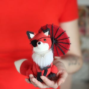 May include: A red crocheted fox wearing a black eye patch and a red and black crocheted umbrella. The fox is holding a white fur collar with a rhinestone pendant.