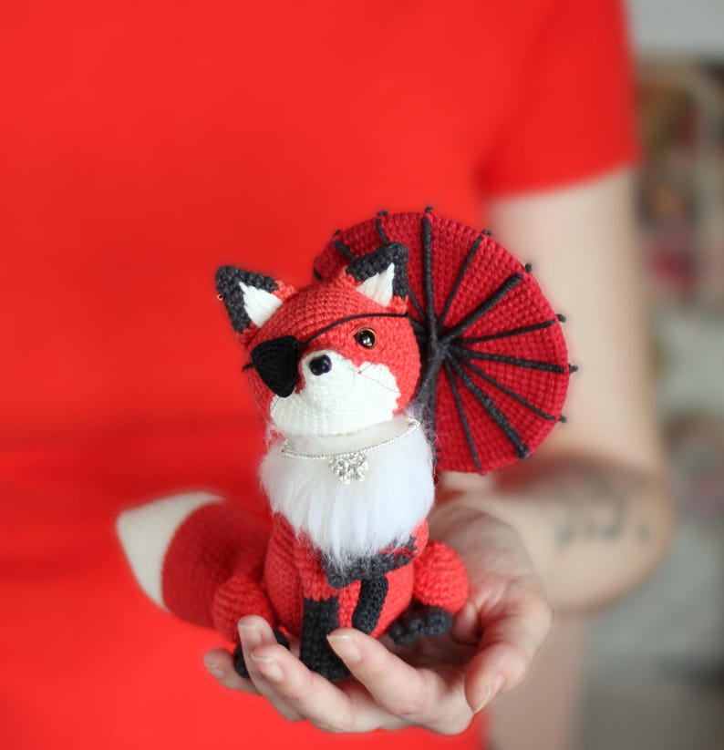 May include: A red crocheted fox wearing a black eye patch and a red and black crocheted umbrella. The fox is holding a white fur collar with a rhinestone pendant.