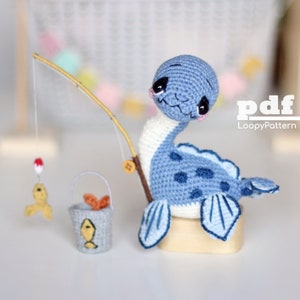 May include: A blue and white crocheted Nessie monster with a fishing rod, a small bucket, and a fish-shaped lure. The Nessie has black eyes and a smiling face. The bucket has a fish design and contains orange objects. The text "pdf LoopyPattern" is visible.