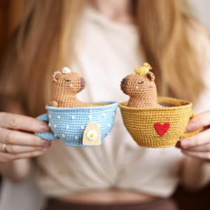 May include: Two crocheted capybaras in teacups. One capybara is in a blue and white polka dot teacup with a daisy tea bag. The other capybara is in a yellow teacup with a red heart.
