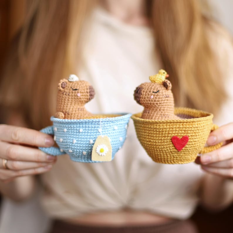 May include: Two crocheted capybaras in teacups. One capybara is in a blue and white polka dot teacup with a daisy tea bag. The other capybara is in a yellow teacup with a red heart.