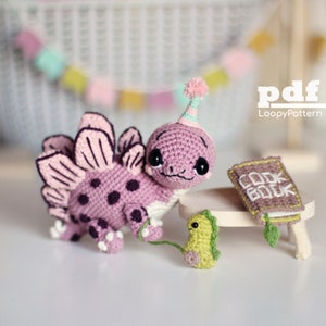 May include: A pink crocheted dinosaur wearing a party hat and holding a green crocheted dinosaur. The dinosaur is standing on a wooden table with a book that says "Cook Book" on it.