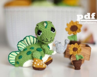 Pode incluir: Um dinossauro de crochê verde segurando um regador cinza, em pé ao lado de um pequeno girassol em vaso. O dinossauro tem olhos pretos grandes e uma barriga branca. O girassol está em um vaso marrom com um caule e folhas verdes. O fundo é uma imagem desfocada de uma cesta e um girassol.