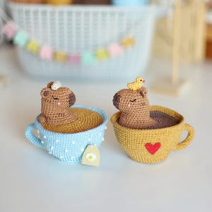 May include: Two crocheted capybaras are relaxing in teacups. One capybara is in a blue teacup with a white daisy tea bag. The other capybara is in a yellow teacup with a red heart. A small yellow crocheted bird sits on the head of the capybara in the yellow teacup.