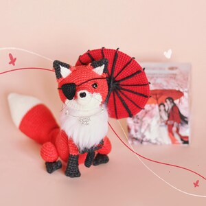 May include: A red crocheted fox wearing an eye patch and a red crocheted umbrella. The fox is sitting on a pink surface.