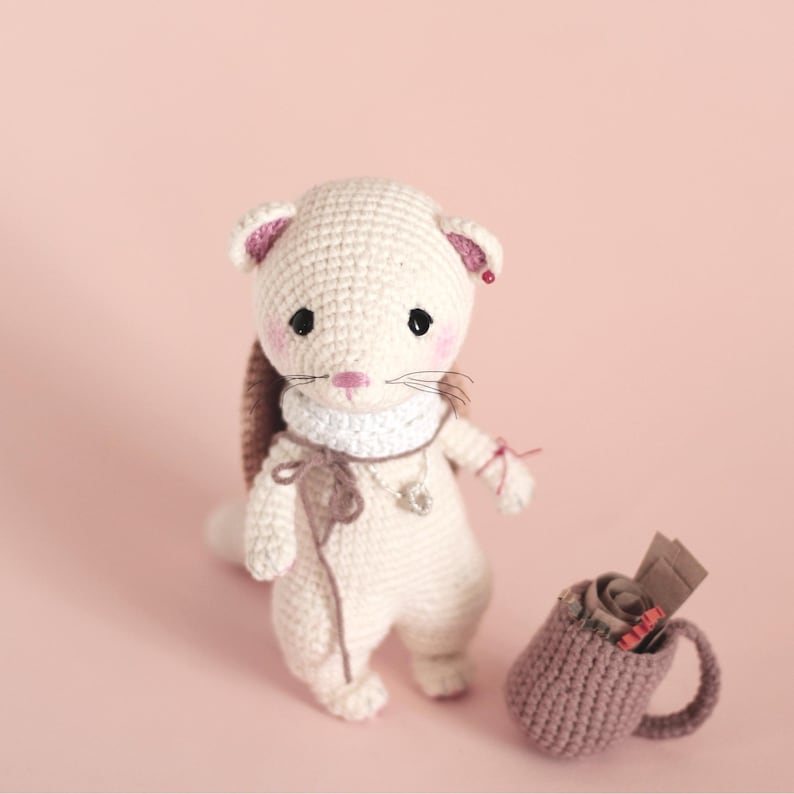 Op de afbeelding: Een wit gehaakt fretje met een witte sjaal en een bruin gehaakt vest. Het fretje staat op een roze oppervlak met een bruin gehaakte mok ernaast.